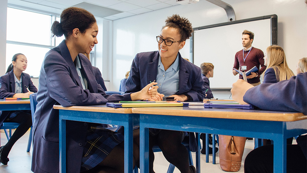 2 teenagers sitting together in a classroom They are smiling and chatting.
